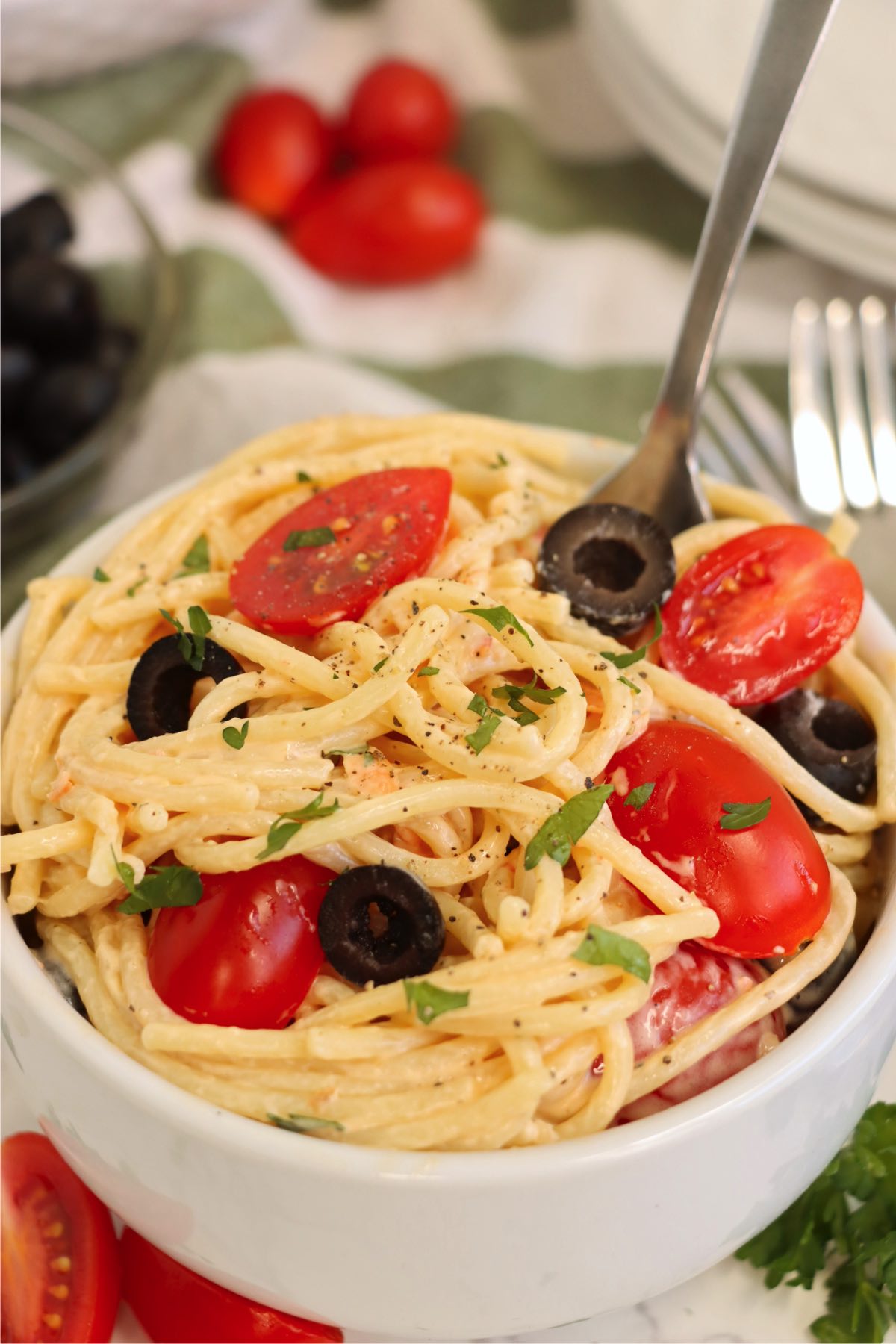 Fork in a small bowl of angel hair pasta salad with black olives and tomatoes.
